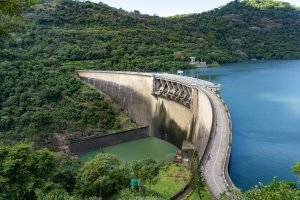 Aerial view of a large dam surrounded by lush green hills and a blue reservoir under a clear sky.